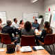 A group of professionals engages in a meeting, discussing charts and plans on whiteboards, with notes and a coffee on the table.